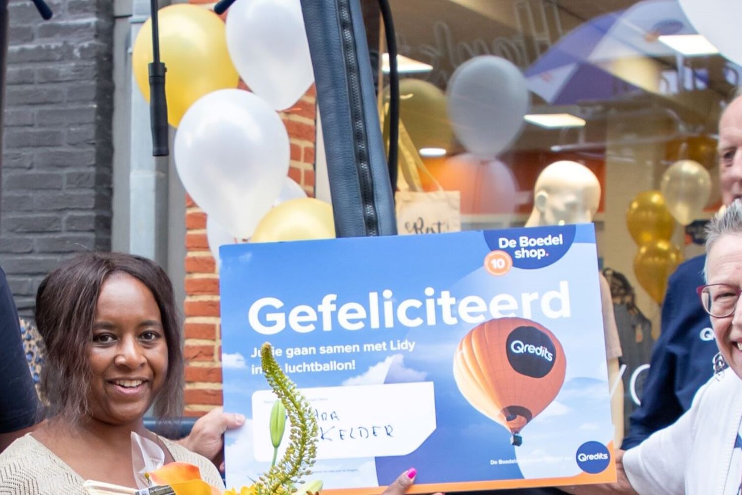 Twee vrouwen staan voor een luchtballon. Iemand heeft een bos bloemen vast en iemand anders een grote kaart.