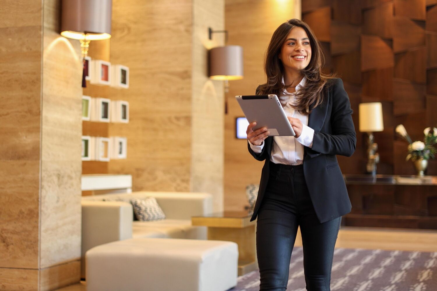 Een lachende vrouw met een pak aan en tablet in haar handen loopt in een lobby. 
