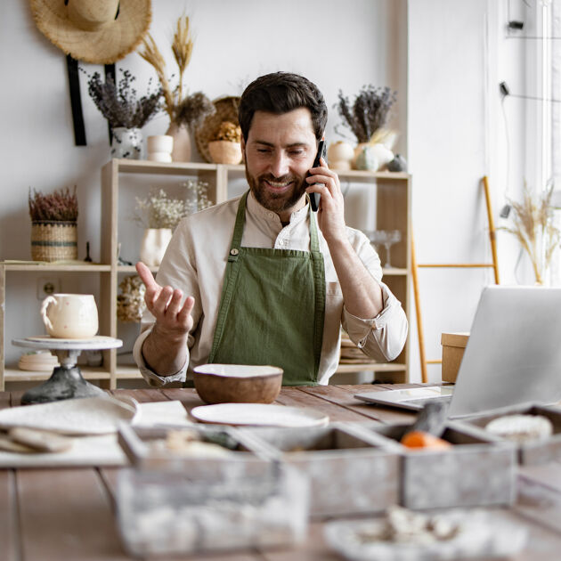 Man met schort zit aan een rommelig bureau en is aan het bellen, hij maakt een handbeweging. 