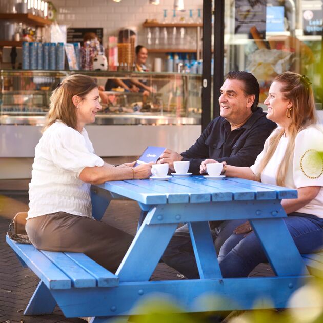 Man en vrouw in gesprek met vrouw aan een blauwe picknicktafel voor een ijssalon. 