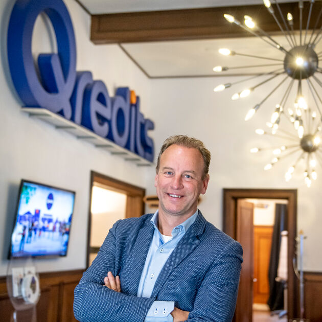 Man in blauw pak staat met zijn handen gevouwen en lacht naar de camera. Hij is in een lichte kamer met Qredits groot geschreven links boven