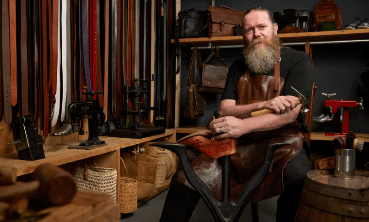 Man met baard en leren schort werkt aan het maken van schoenen en kijkt naar de camera