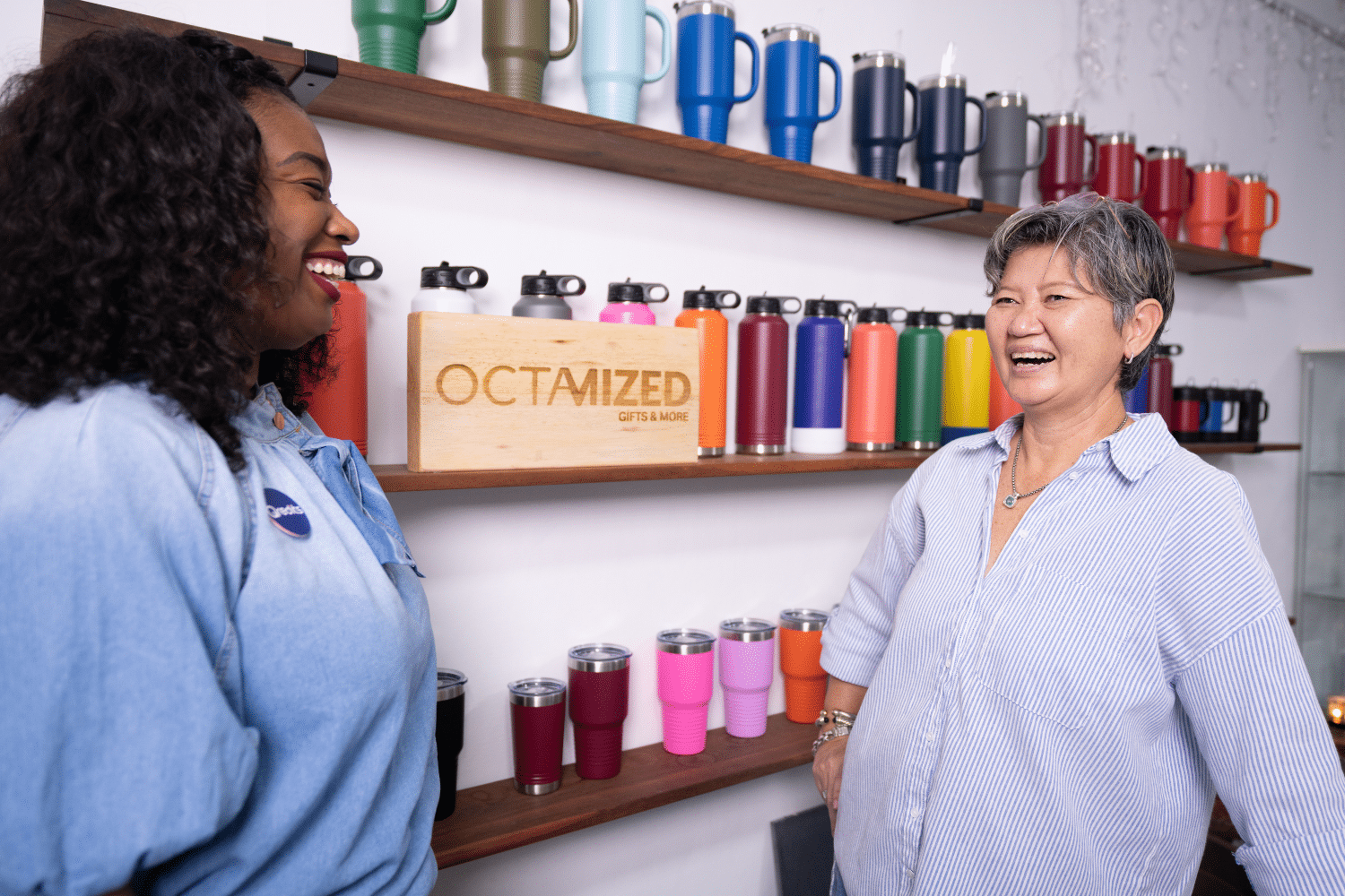 Twee vrouwen staan voor een rek thermoflessen en lachen naar elkaar.