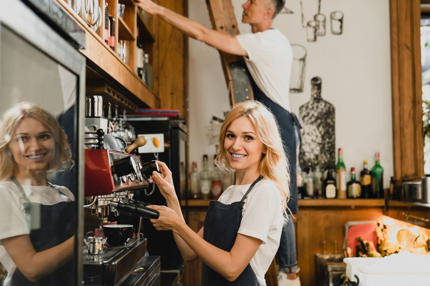 Vrouw zet koffie bij het koffiezetapparaat, man in de achtergrond staat op de ladder.