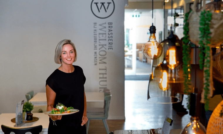 Vrouw in zwarte kleding staat voor een witte muur in een restaurant. Ze heeft eten vast