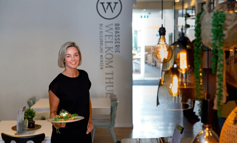 Vrouw in zwarte kleding staat voor een witte muur in een restaurant. Ze heeft eten vast