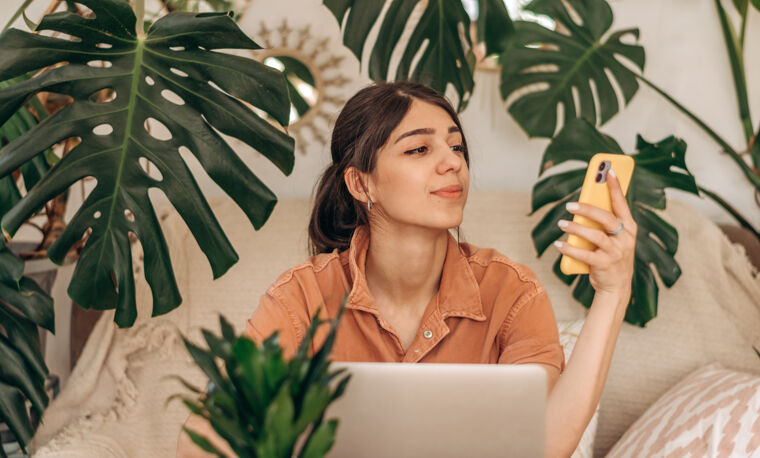 Vrouw zit achter de laptop en kijkt naar haar telefoon met veel planten