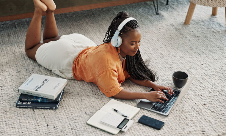 Vrouw ligt op de grond te werken op de laptop met een koptelefoon op. 