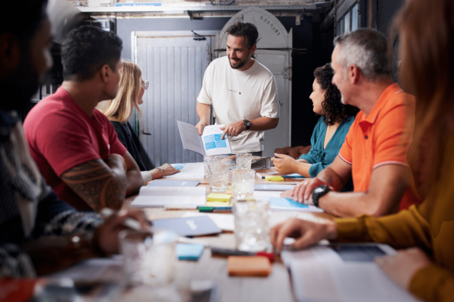 Man in wit shirt geeft les aan tafel vol mensen 