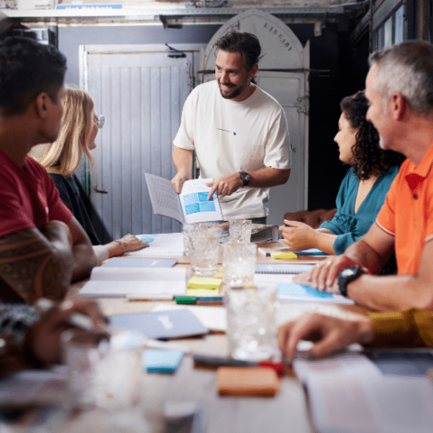 Man in wit shirt geeft les aan tafel vol mensen