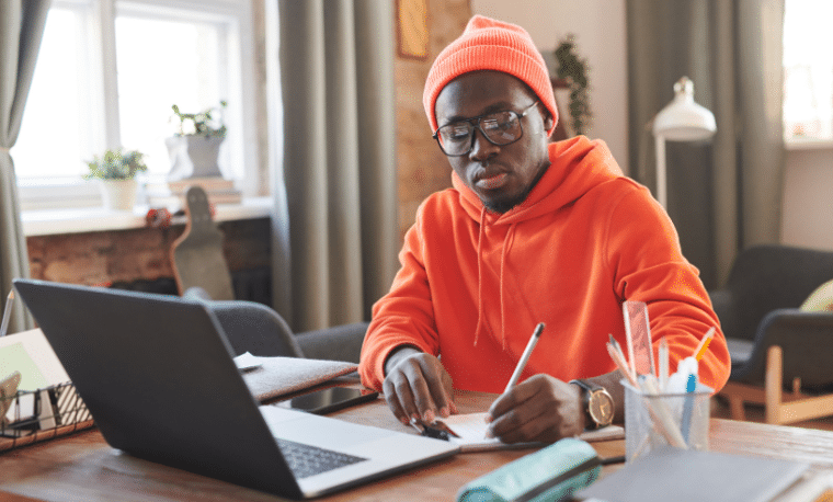 Man in oranje sweater en muts zit achter zijn laptop te schrijven