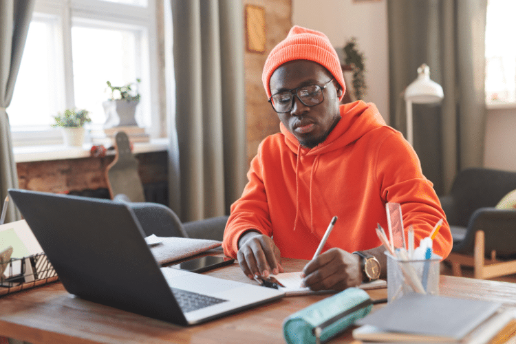 Man in oranje sweater en muts zit achter zijn laptop te schrijven
