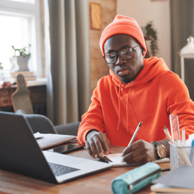 Man in oranje sweater en muts zit achter zijn laptop te schrijven