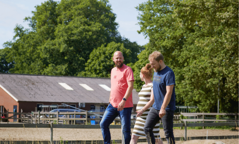 drie mensen lopen in een weiland met een boerderij in de achtergrond