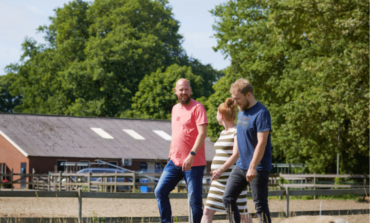 drie mensen lopen in een weiland met een boerderij in de achtergrond