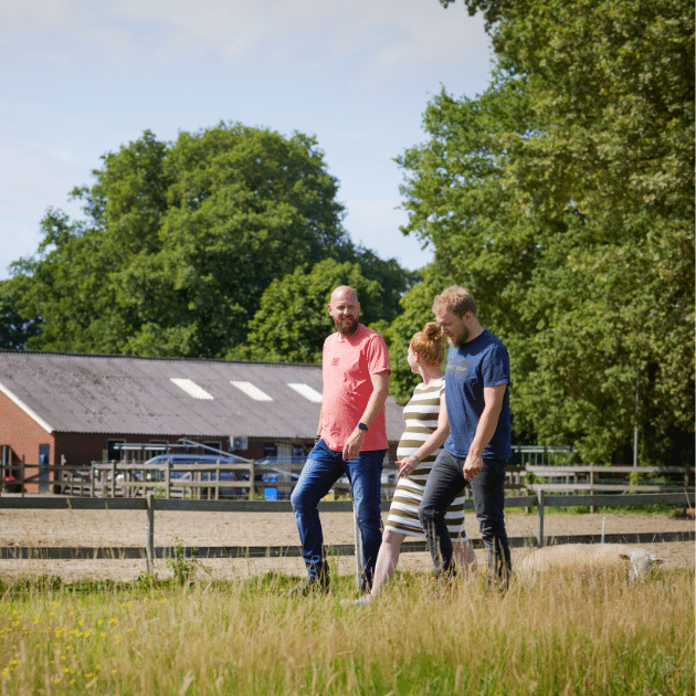drie mensen lopen in een weiland met een boerderij in de achtergrond