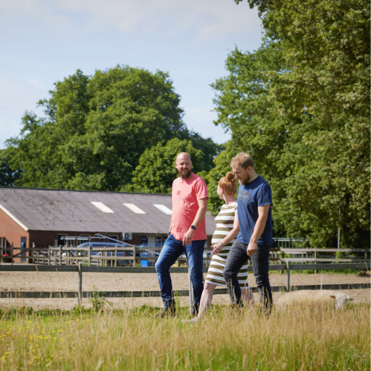 drie mensen lopen in een weiland met een boerderij in de achtergrond
