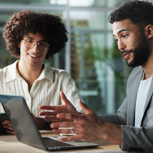 Twee mannen zitten aan bureau en kijken opgetogen naar laptop.