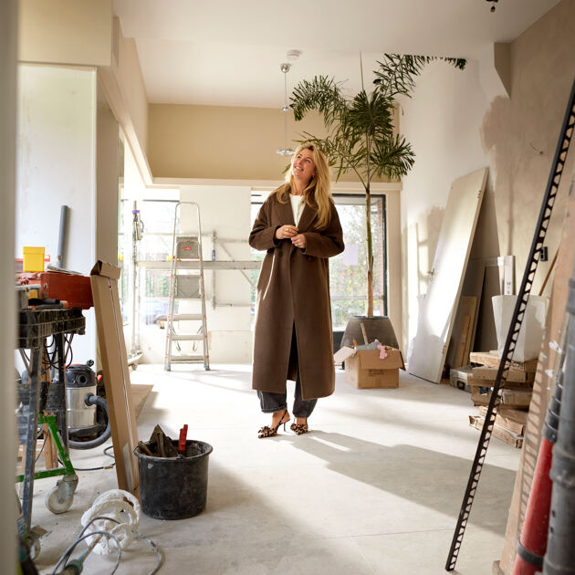 Vrouw staat middenin verbouwing, met hakken en een lange bruine jas aan.