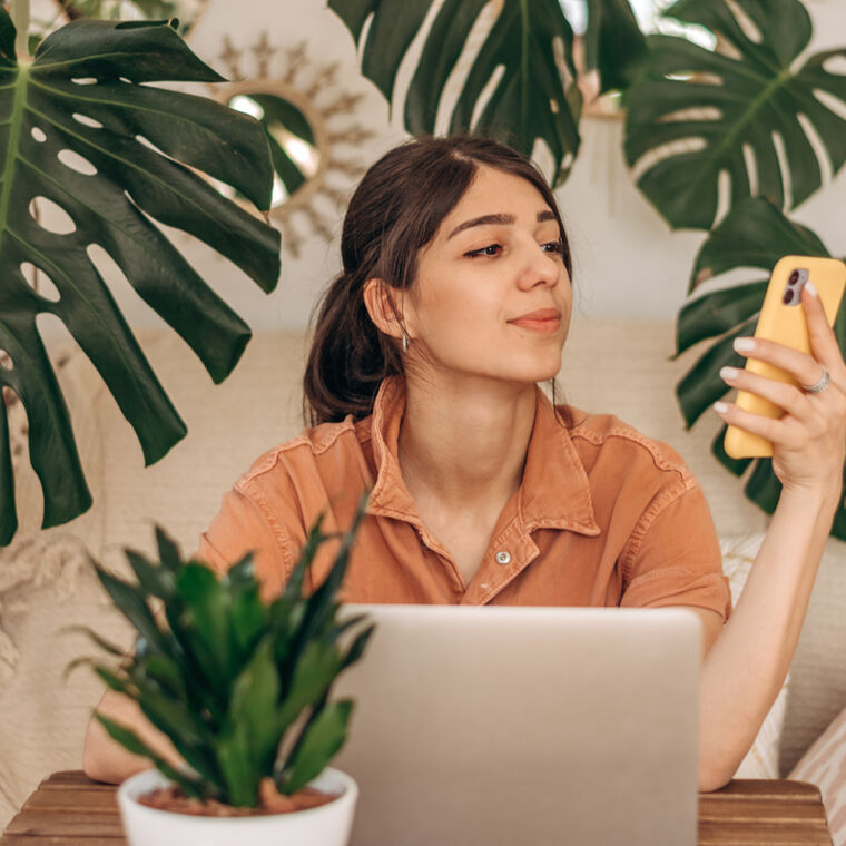 Vrouw zit achter de laptop en kijkt naar haar telefoon met veel planten