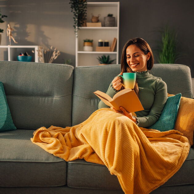 Vrouw zit lachend op de bank, ze leest een boek en heeft een mok in haar hand.