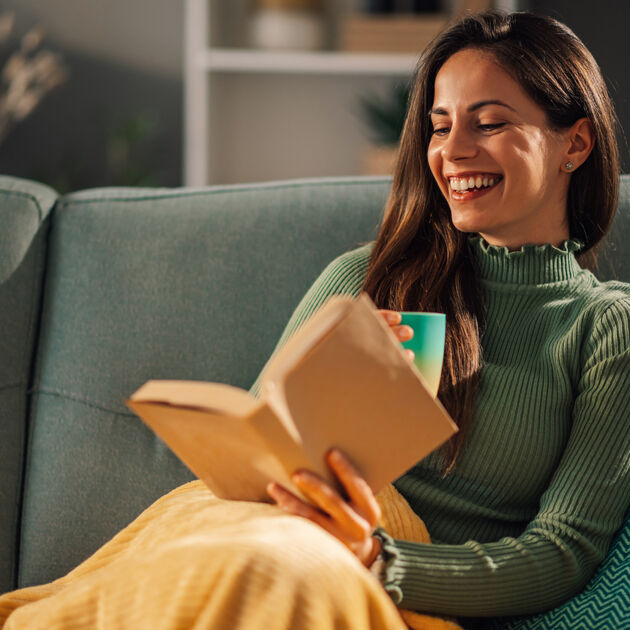 Vrouw zit op de bank en lacht, ze heeft een boek in haar hand.