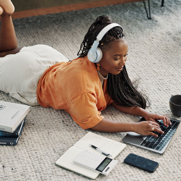 Vrouw ligt op de grond te werken op de laptop met een koptelefoon op. 