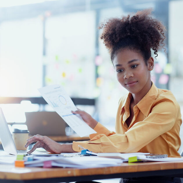 Vrouw met gele blouse met papieren in de hand kijkt naar laptop. 