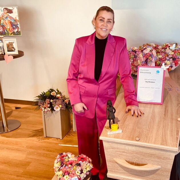 Vrouw met roze pak staat naast een rouwkist.