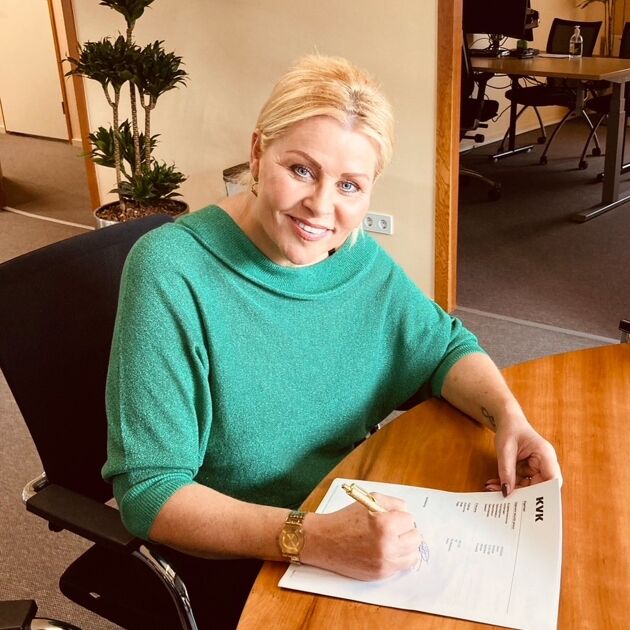 Vrouw zit aan tafel, kijkt lachend en tekent papier. 
