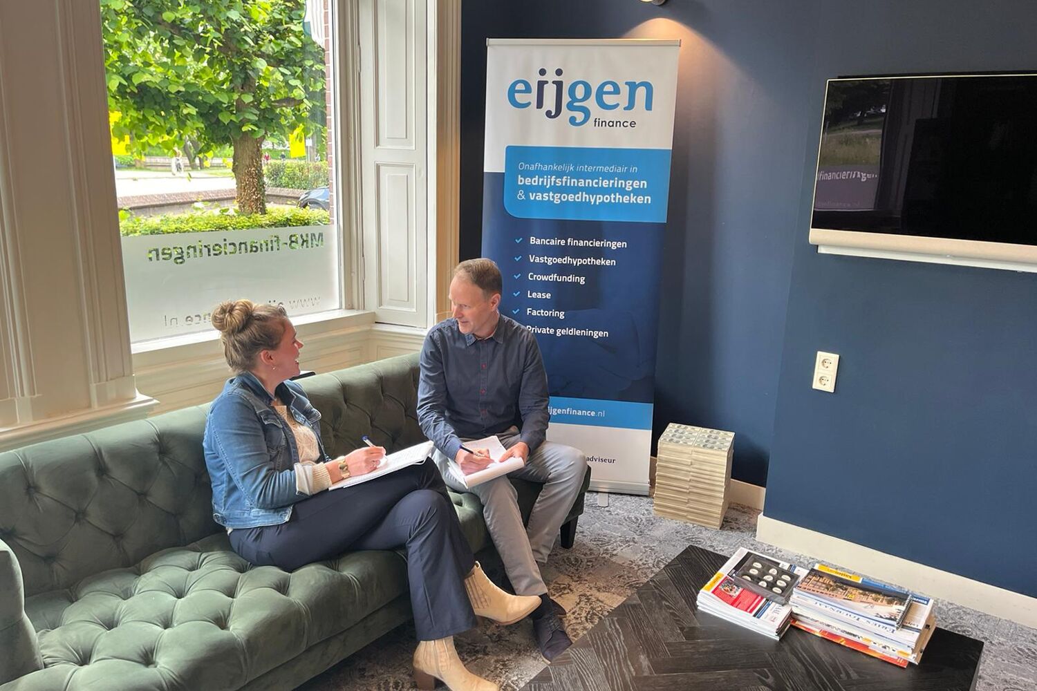 Vrouw in spijker jasje en man in blauw shirt zitten op groene bank bij raam