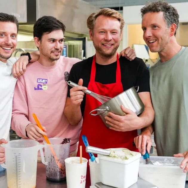 Vier mannen staan lachend in de keuken. Een heeft een rood schort en garde. Een heeft een groen shirt. Een heeft een roze shirt en een spathula. Een heeft een wit shirt en kijkt naar de camera