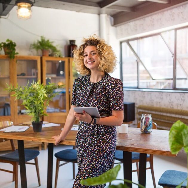 Een vrouw met krullend haar leunt tegen een bureau met een ipad in haar handen. 