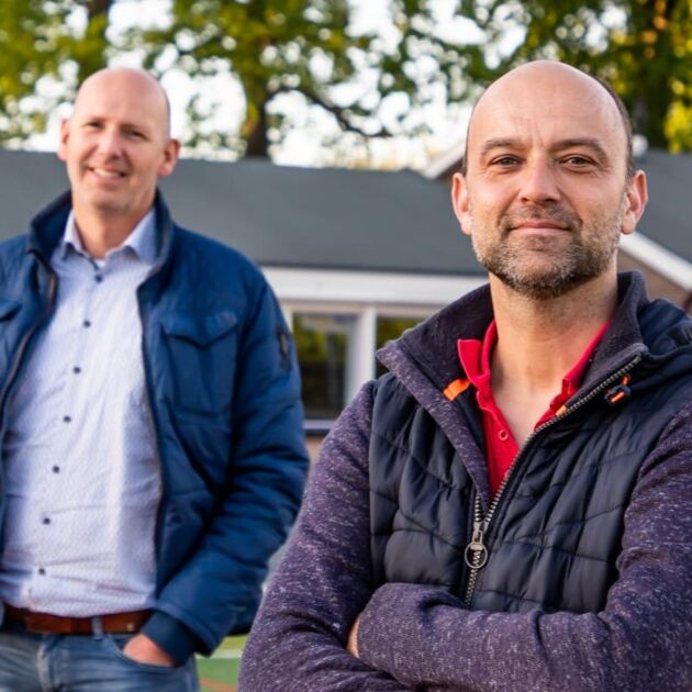 Twee mannen staan buiten op een sportveld. Ze lachen beide naar de camera