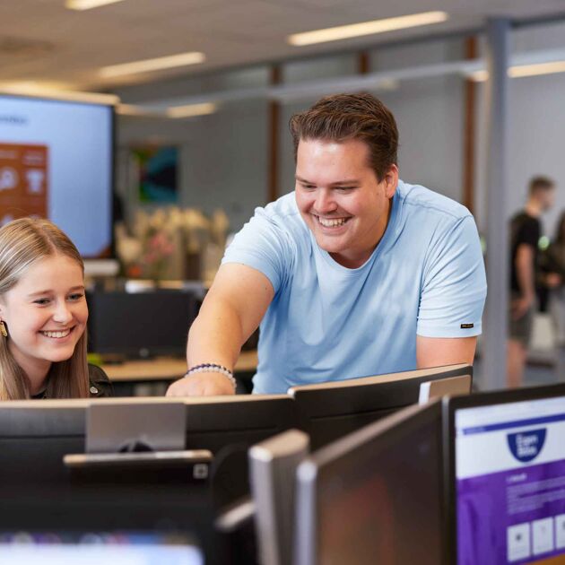 Man met blauw shirt zit naast student en wijst naar computerscherm