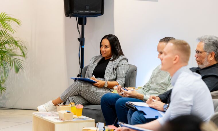 Een vrouw in pak zit op een grijze stoel terwijl zij dingen opschrijft. Er zitten twee mannen naast haar