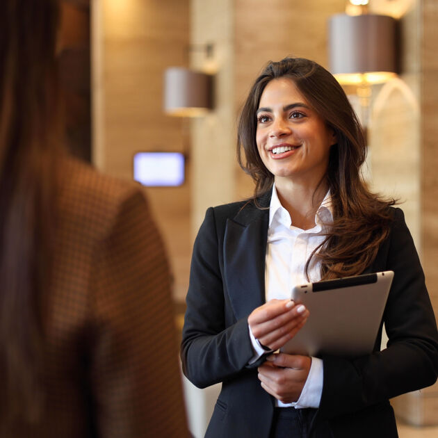 Een vrouw met een pak aan en tablet in haar handen kijkt lachend naar een andere vrouw. 
