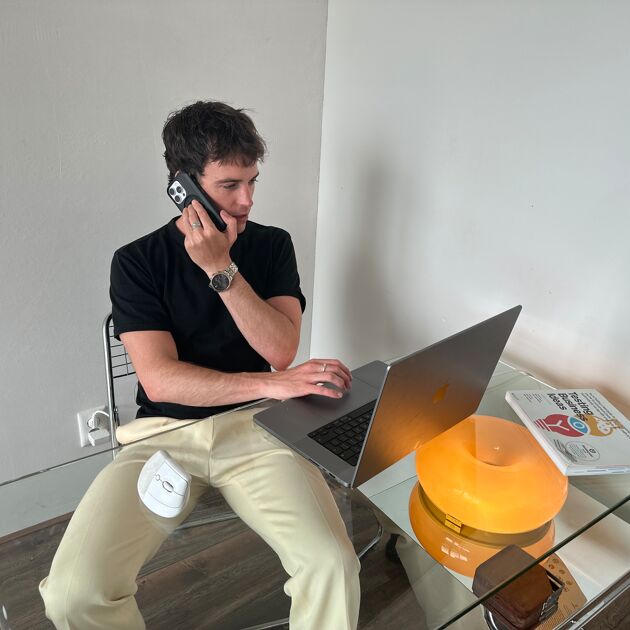 Een man zit achter een bureau en is aan het bellen, hij kijkt op zijn laptop. 