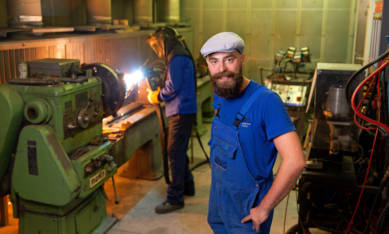 Man in een werkplaats in blauwe overalls, lasser op de achtergrond