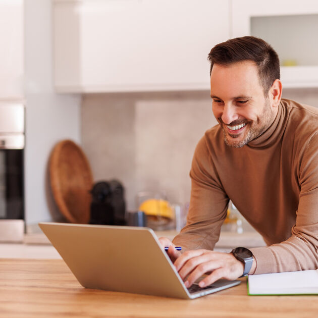 Lachende man leunt op het aanrecht en kijkt naar laptop. 