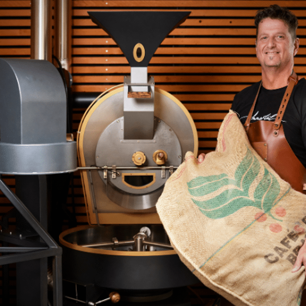 Man in een leren schort heeft een grote jute zak met koffiebonen vast en staat voor een grote machine.