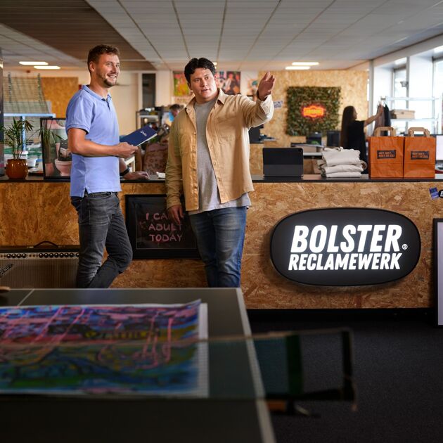 Twee mannen staan voor een houten balie met logo bolster reclamewerk. 