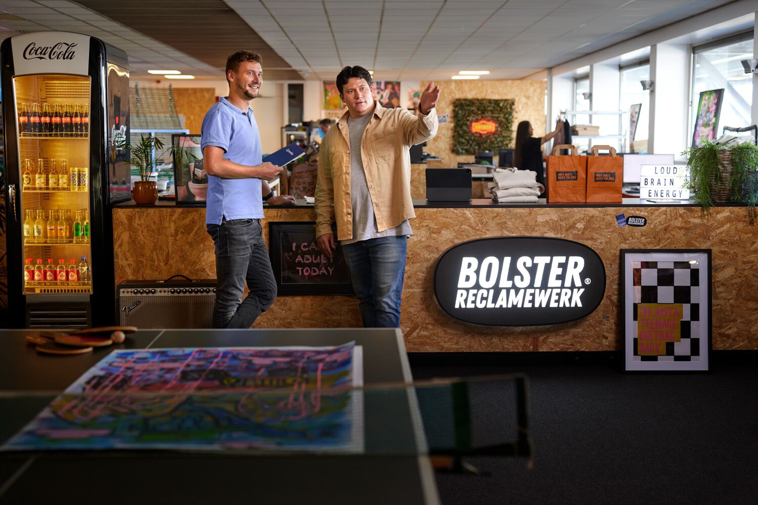 Twee mannen staan voor een houten balie met logo bolster reclamewerk. 