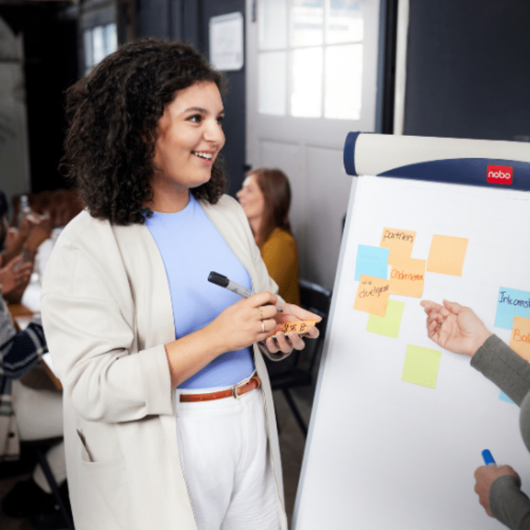 Vrouw met blauw shirt en grijs jasje schrijft iets op een post-it voor op een whiteboard