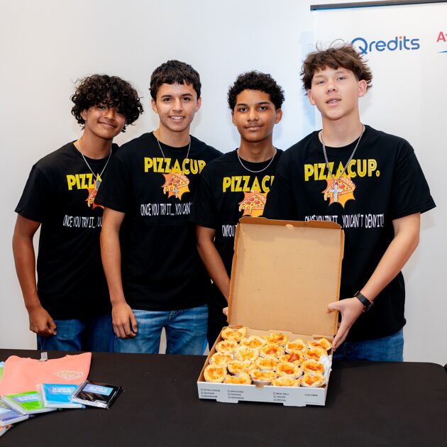 Vier jongens in zwarte shirts staan achter een zwarte tafel met een kartonnen pizzadoos die open is