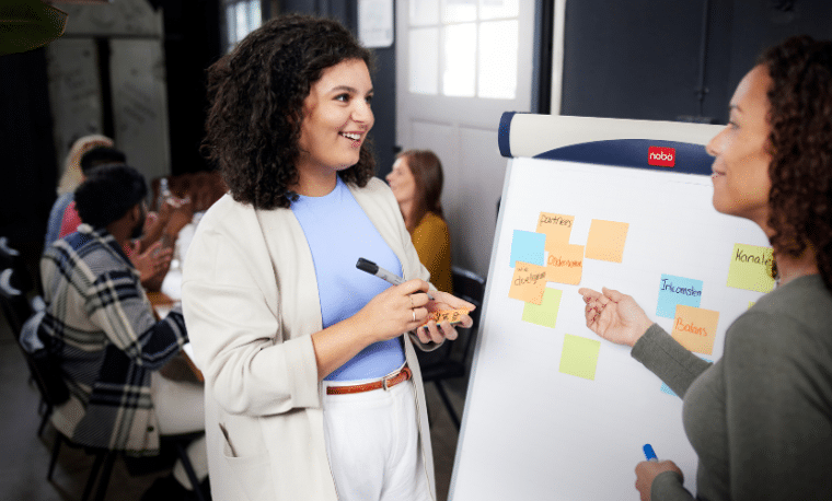 Vrouw me blauw shirt en grijs jasje schrijft iets op een post-it voor op een whiteboard