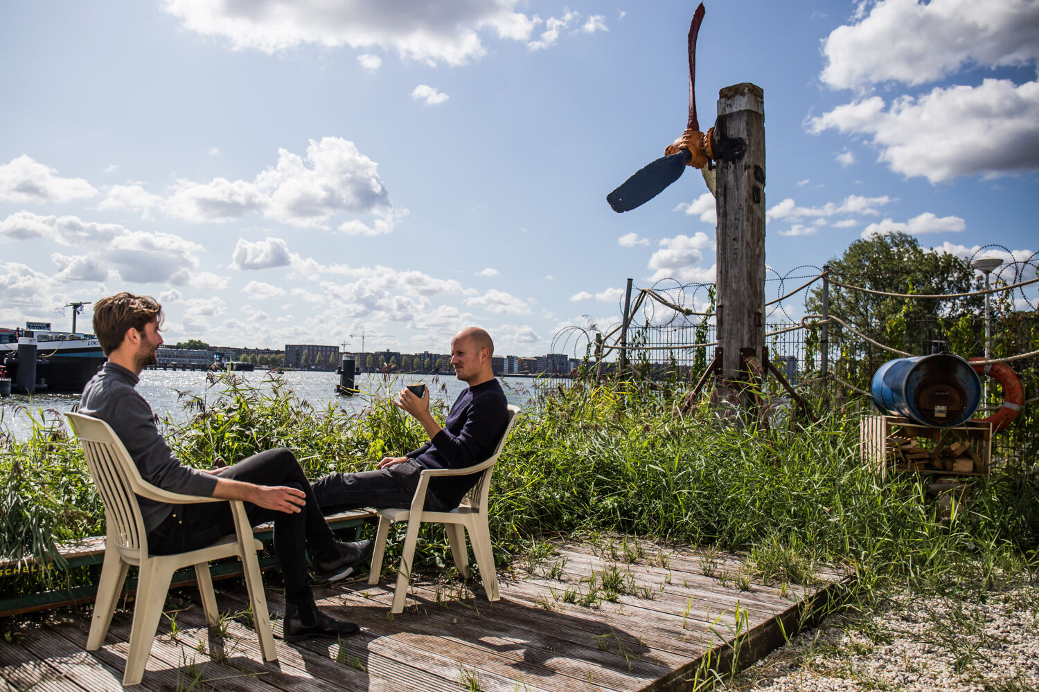 Twee mannen zitten aan het water op houten stoelen met veel groen omheen