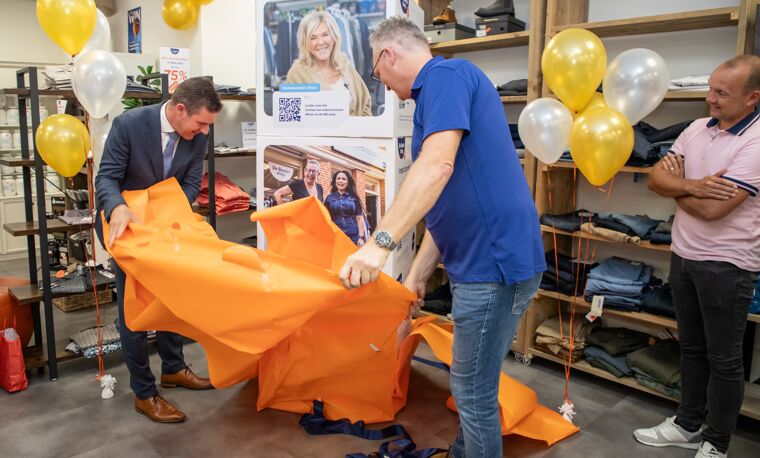 Twee mannen scheuren oranje papier weg van een stapel blokken. Er zijn gele en witte balonnen.