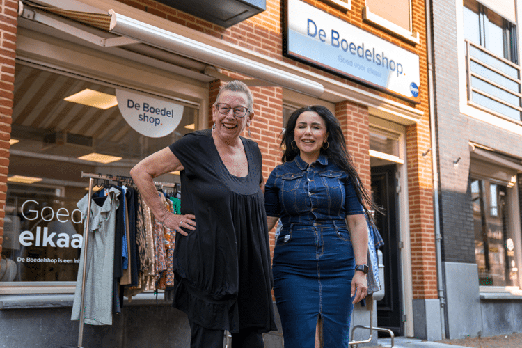 Twee vrouwen staan voor de boedelshop en lachen naar de camera