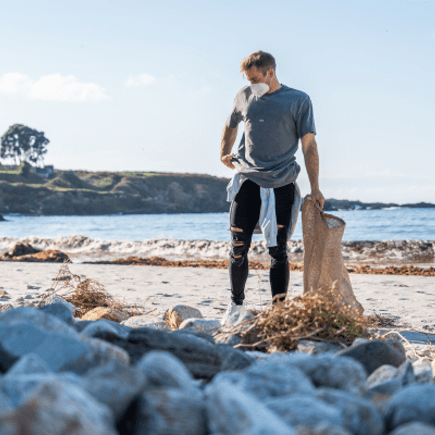 man loopt op het strand en is op zoek naar afval, hij heeft een zak met afval vast
